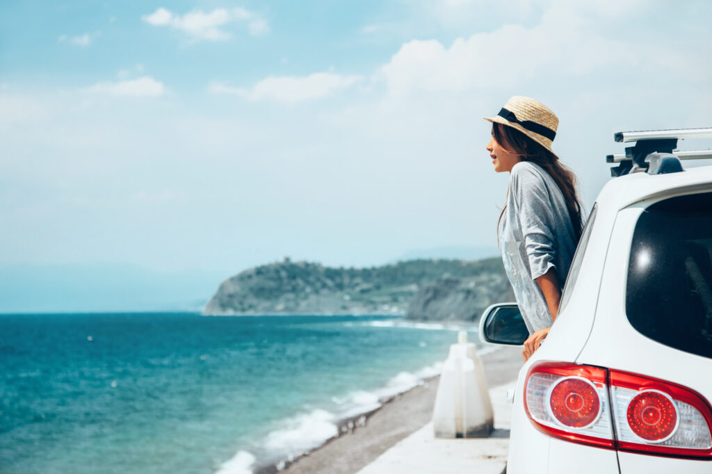 woman wearing hat looking out of car window towards blue ocean for travel