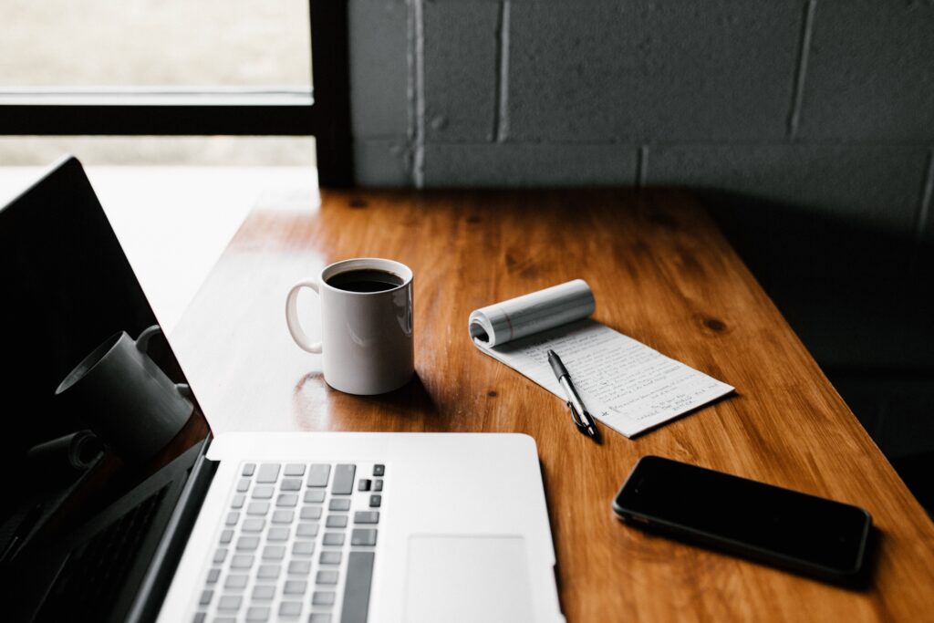 open laptop, coffee mug, phone, notepad and pen on a wooden desk