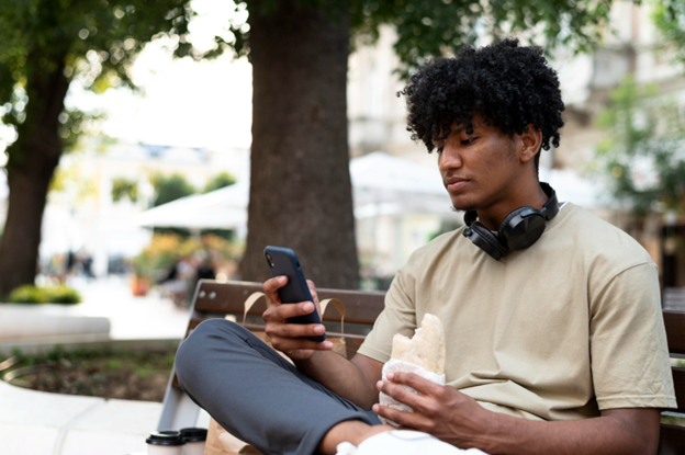 young man on park bench holding a sandwich and looking at his mobile phone